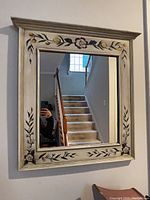 Front view of the square mirror mounted on a wall, showing the hand painted floral design on the cream colored wooden frame.