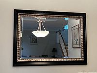 Mirror mounted on a wall above a dining table with black chairs and a chandelier above, showing the frame and clear reflective surface.