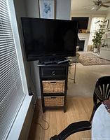 Photo showing flat-screen TV on top of a black TV stand with two woven baskets below and a drawer above the baskets.