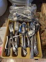 Tray with assorted used metal forks, spoons, and knives laid out inside a beige plastic utensil organizer with additional utensils in a clear plastic bag on top.