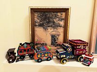 Photo showing six colorful, hand-painted wooden toy vehicles lined up on a shelf with a framed print behind them.