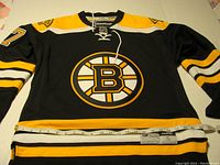 Front full view of black Boston Bruins jersey with yellow and white stripes, centered Bruins logo, and #37 on sleeve with measuring tape showing size.