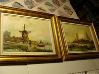 Pair of vintage Dutch landscape oil paintings in gold-tone wooden frames, showing rural scenes with windmills, houses, boats, and trees.