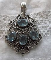 Front view showing ornate open-work sterling silver pendant with four oval faceted light blue stones set in bezel mounts, detailed bail on top