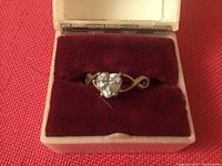 Front view of sterling silver engagement style ring with heart-shaped stone inside cream-colored box with dark red velvet interior.