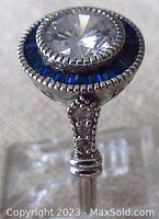 Close-up of top of the ring showing clear round facetted center stone surrounded by keystone-shaped blue stones in channel setting.