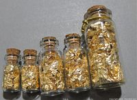 Front view of five graduated glass bottles filled with gold foil flakes, cork sealed, largest bottle with gold chain