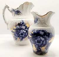 Two antique ceramic wash set pieces: toothbrush holder and small pitcher with flow blue floral and gold decoration, front view