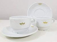 Two white porcelain mugs and saucers with gold crown monogram shown together on a white surface
