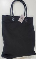 Front view of a black canvas tote with two black glossy leather-like handles and an attached NB Nobia tag, showing the bag's general shape and texture.