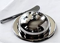 Polished stainless steel butter dish with lid on, and stainless steel spreader placed beside it on white surface.