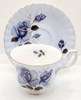 Cup and saucer set with blue rose design and gold trim. Cup front view showing floral pattern and gold rim. Saucer in background with scalloped gold rim and floral decoration.