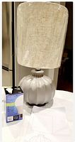 Gray finished ribbed rounded base lamp with beige textured drum shade next to LED bulb packaging on white surface