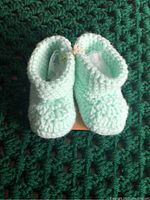 Pair of mint green crocheted baby booties placed on a green textured surface, showing the front and top view.
