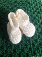 Pair of white handcrafted baby booties on a green crocheted blanket background