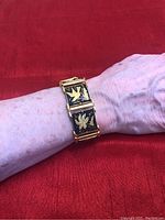 The bracelet is shown worn on a wrist against a red fabric background. The bracelet has hinged gold toned segments decorated with black panels featuring intricate gold bird motifs and a clasp closure.