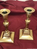 Photo of two metal candlesticks on a maroon fabric background showing square bases, tapered columns, and round candle holders with worn patina.