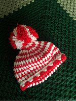 Front view of a handcrafted red and white crocheted toque with a pom-pom and a ripped-edge brim design on a green textured background.