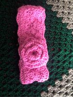 Close-up photo showing a bright hot pink, handcrafted crocheted headband with a rose-like flower on top, placed on a textured fabric background.