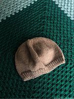 Photo of beige, handcrafted knitted toque laid flat on a green and white crocheted blanket.