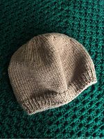 Beige knitted toque hat laid on a green textured surface showing detailed knitting stitches and ribbing at the bottom edge.