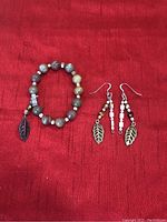Image showing a handcrafted beaded bracelet and a pair of matching dangle earrings laid on a red fabric background, highlighting the natural stone beads and metal leaf charms.