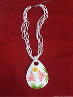 Photo of white multi-strand beaded necklace with teardrop-shaped shell pendant painted with pink and yellow flowers and green leaves, laid on a red fabric background.