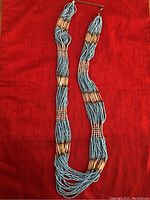Long beaded necklace shown laid flat on red fabric background, displaying the pattern of blue and tan beads and metal clasp