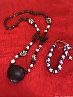 Photo showing handcrafted necklace and matching bracelet on red fabric background. Necklace has diverse bead types including carved and white beads; bracelet has rounded purple beads.