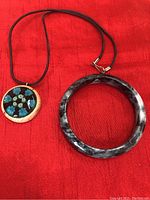 Photo of a pendant necklace with a floral design and a black cord next to a marbled black and gray bangle-style bracelet on a red cloth background.