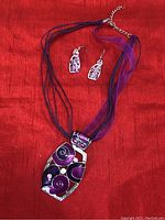 Photo showing handcrafted necklace with multi-strand purple chains and large pendant, alongside matching purple and silver earrings, set on a red background.