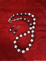 Photo of handcrafted necklace with alternating large silver and black round beads paired with matching earrings with similar beads on a red cloth background.