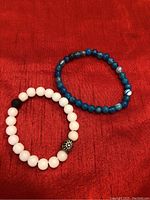 Photo of two handcrafted beaded bracelets on a red fabric background, one white with decorative metal bead and one blue.