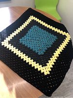 Full view of small crocheted blanket folded on table showing square color pattern in black, yellow, and teal