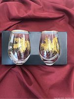 Pair of clear stemless wine glasses with gold decorative leaf designs displayed on black box and burgundy fabric background.
