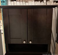 Front view of the closed 25" espresso medicine cabinet showing double paneled doors with silver round knobs and an open bottom shelf.