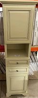 Closed view of antique white linen tower with doors, drawers, and open middle shelf.