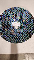 Top view of round glass vessel sink showing colorful checkered mosaic pattern in blue, green, brown, and black small square tiles with central drain hole.