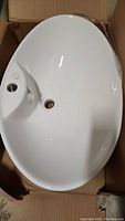 Oval white ceramic vessel sink with faucet and drain holes visible, placed inside cardboard box
