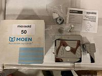 Moen single handle shower trim kit components including chrome handle, escutcheon, cartridge, and mounting hardware on white surface beside the original Moen box labeled with MaxSold.