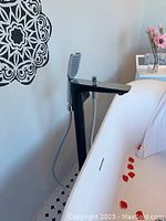 Black freestanding tub faucet installed beside a white bathtub with petals inside. Faucet has a flat spout and attached hand shower with hose.