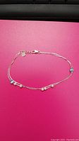 Sterling silver ankle bracelet with small beads shown on a pink background in oval shape with lobster clasp and charm.