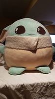Front view of large Baby Yoda plush toy sitting on bed, showing detailed facial embroidery, ears, and beige collar.