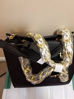 Front and top view of black handbag showing its structured shape, chain handles wrapped in plastic and original tags.