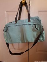 Front view of a light blue duffle bag hanging from a door handle, showing top zipper and dark gray straps.