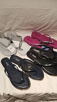 Four pairs of ladies flip flops laid on light-colored fabric, showing different colors and styles including white with silver straps, hot pink, black glossy, and navy.