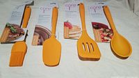 Photo of 4 orange silicone kitchen utensils laid out including basting brush, spatula, slotted turner, and spoon. Original packaging tags visible.