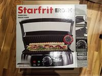 Front view of the Starfrit Panini Grill box showing the grill and a pressed sandwich, highlighting removable plates, adjustable temperature control, and extra thick ridged plates.