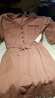Photo of pink DeX dress laid out on surface, showing front buttons, chest pockets, and fabric belt with tag attached