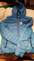 Front view of light blue ladies medium puffer jacket laid flat showing hood, full zipper with black trim, and long sleeves.